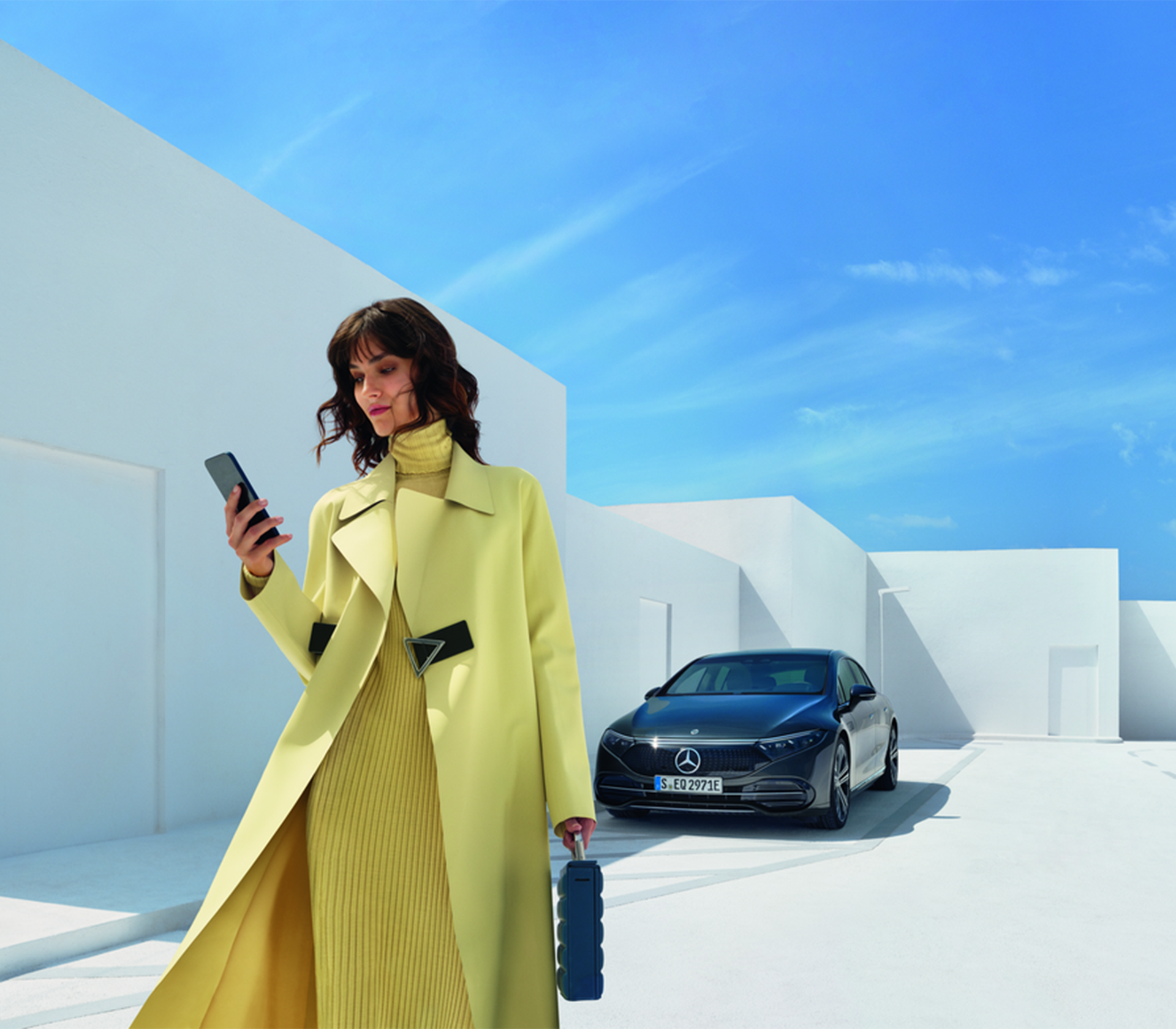 Eine Frau in einem gelben Mantel und Kleid läuft und schaut auf ihr Handy während ihr Auto im Hintergrund geparkt ist